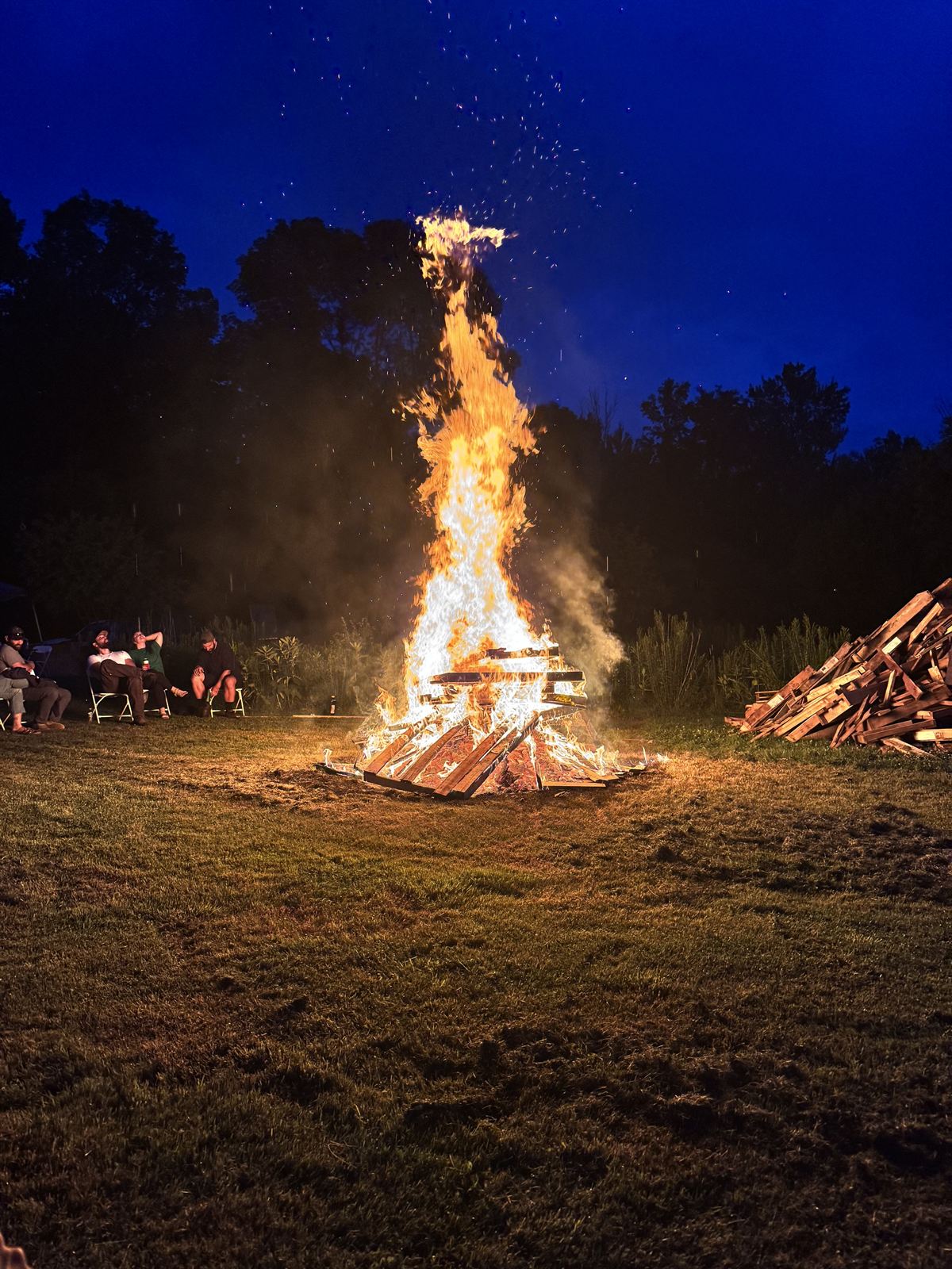 The bonfire fully lit at dusk with people in camp chairs around it